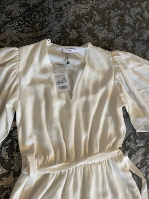 Vit långklänning med puffärm  - Elegant vit långklänning  med v-ringning, puffiga halvtransparenta ärmar och knytband i midjan. Klänningen har en slits på sidan och är tillverkad i ett lätt, mjukt material som ger ett flowy intryck. Perfekt för sommarens alla tillfällen. Aldrig använts innan och storlek S jätte fin inför studenten och allmänt 