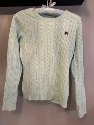 Ljusgrön kabelstickad tröja Bondelid - Snygg ljusgrön kabelstickad tröja från Bondelid med rund halsringning och diskret logga på bröstet. Tröjan har långa ärmar och ribbade muddar vid ärmslut och nederkant. Perfekt för dig som gillar klassisk och stilren design.