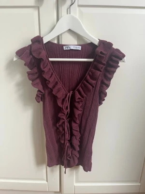 Vinröd ribbad linnetopp från ZARA - Trendig vinröd linnetopp från ZARA med volangkant längs v-ringningen och snörning framtill. Toppen är ribbad och ärmlös, perfekt för att lyfta vilken outfit som helst med sin unika design och snygga detaljer.