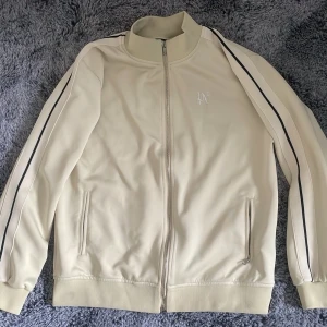 Palm angels kofta  - Snygg beige ziptröja med svarta ränder längs ärmarna och diskret logga på bröstet. Tröjan har hög krage, dragkedja framtill och två fickor med dragkedja. Perfekt för en sportig och avslappnad stil. 