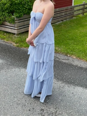 Ljusblå långklänning med volanger - Säljer en ljusblå långklänning med flera lager volanger och axelbandslös design. 💕🌸Klänningen har en stor rosett framtill och är tillverkad i ett tunt, flygigt material som ger en romantisk känsla. Det går även att ta bort rosetten om man hellre vill ha den som scarf 💕🌸Perfekt för bal, bröllop eller bara en fin sommardag☀️🌸💕
