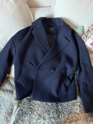 Mörkblå kort kappa från Lindex - Stilren mörkblå kappa från Lindex med dubbelknäppning och bred krage. Modellen är kortare och har två snedställda fickor framtill. Materialet är tjockt och känns ulligt, perfekt för kyligare dagar. Enkel knappdetalj framtill för en clean look. Den har använts fåtal gånger. Nypris 1000kr. Storleken är XS men det är en oversized passform 💕