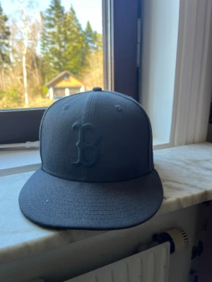 Mörkgrå Boston keps från New Era - Säljer en mörkgrå keps från New Era med Boston B-logga broderad framtill. Klassisk flat brim och strukturerad form, perfekt för dig som gillar sportiga accessoarer. Materialet är slitstark bomull och kepsen har en stilren look.