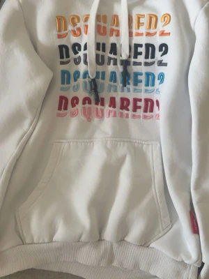 Vit DSQUARED2 hoodie med brodyr - Snygg vit hoodie från DSQUARED2 med broderad logga i flera färger på bröstet. Hoodien har huva med dragsko och en stor magficka. Tillverkad i mjukt bomullsmaterial som känns skönt mot huden. Perfekt för dig som gillar streetwear och vill sticka ut.
