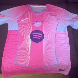 Rosa Barcelona fotbollströja spelarversion  - Snygg Barcelona fotbollströja från Nike i rosa och ljusblått med vita grafiska linjer och klubbmärke på bröstet. Tröjan har korta ärmar och är tillverkad i ett lätt, ventilerande material. Spotify-logga på framsidan och modern design som sticker ut på planen.