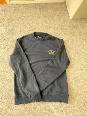 Mörkblå sweatshirt från Bondelid - Mörkblå sweatshirt från Bondelid med tryck på bröstet där det står 'Bondelid Athletics EST 1956'. Klassisk rund halsringning och ribbade muddar vid ärmslut och nederkant. Perfekt för en avslappnad och sportig stil.