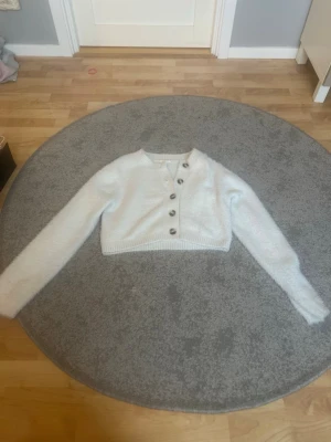 Vit fluffig kofta med knappar - Snygg vit kofta med fluffigt material och stora, mörka knappar framtill. Kort modell med långa ärmar och ribbstickade detaljer vid nederkant och ärmslut. Perfekt för att skapa en trendig och mysig look💓💓