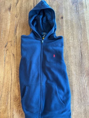 Mörkblå hoodie från Polo Ralph Lauren - Säljer en mörkblå hoodie från Polo Ralph Lauren med dragkedja och huva. Tröjan har två fickor framtill och ett litet rött Polo-logo broderat på bröstet. Materialet är mjukt och bekvämt, perfekt för chill dagar. Klassisk och stilren design.