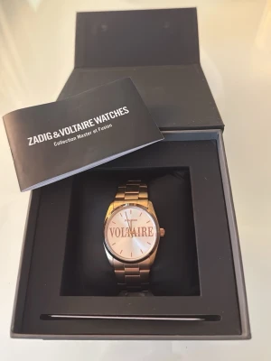 Guldfärgad klocka från Zadig & Voltaire - Snygg guldfärgad klocka från Zadig & Voltaire med metallarmband och stilren urtavla där märket står tydligt. Klockan kommer i originalbox med extra länkar och garantikort. Perfekt accessoar för dig som vill ha något trendigt och elegant.