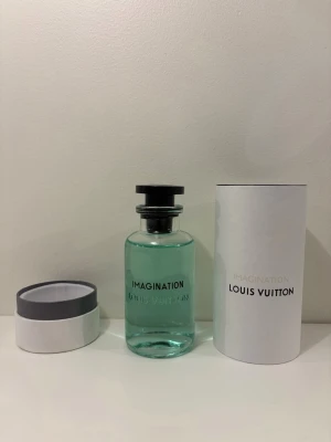 Louis Vuitton Imagination parfym 100ml - Exklusiv parfym från Louis Vuitton, Imagination, i en stilren glasflaska med turkosgrön vätska och svart lock. Medföljer elegant vit cylinderformad förpackning med grå detaljer och tydlig logotyp. Perfekt för dig som vill ha en lyxig doftupplevelse.