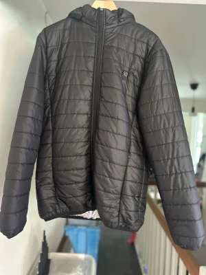Svart pufferjacka med huva uppvärmning  - Stilren svart pufferjacka med huva och dragkedja framtill. Jackan har quiltad design med horisontella sömmar och en liten logotyp på bröstet. Perfekt för kalla dagar och enkel att matcha med olika outfits.Uppvärmd jacka som fungerar med powerbank. Den har tre värmenivåer, och när du inte vill ha värme kan du stänga av den – då fungerar den som en vanlig regnjacka.