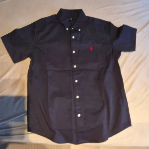 Marinblå kortärmad skjorta Polo Ralph Lauren - Snygg marinblå skjorta från Polo Ralph Lauren med korta ärmar och klassisk krage. Skjortan har vita knappar framtill och en röd broderad logga på bröstet. Perfekt till jeans eller chinos för en clean look.