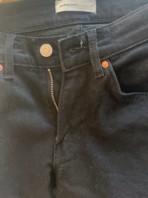 Svarta jeans från Perfect Jeans - Svarta jeans från Perfect Jeans med dragkedja och knapp framtill. Jeansen har slitna detaljer på båda benen och klassisk femficksdesign. Perfekta för en avslappnad och trendig look.