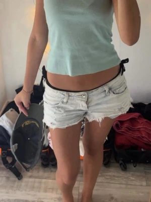Ljusa jeansshorts med slitningar - Säljer ett par ljusblå jeansshorts med råa, fransiga kanter och slitna detaljer. Shortsen har klassisk femficksmodell och knappgylf. Perfekta för en avslappnad och trendig sommarstil.