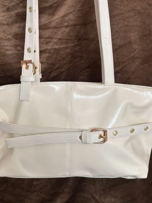Vit handväska med bältesdetalj - Stilren vit handväska i glansigt konstläder med justerbara axelband och snygga guldfärgade spännen. Väskan har en unik bältesdetalj framtill och enkel, modern design som passar till många outfits. Perfekt för dig som gillar minimalistisk och trendig stil.