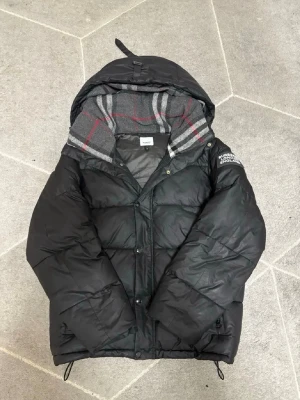 Svart pufferjacka med huva - Säljer en svart pufferjacka från Burberry med stor huva och rutigt foder i grått, rött och vitt. Jackan har dragkedja och tryckknappar framtill samt snörning i nederkant. Märkeslogga på ärmen och klassisk oversized passform.