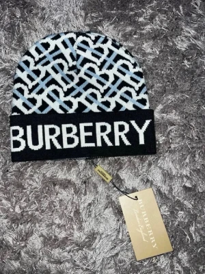 Mössa - Säljer en snygg  mössa med monogrammönster i svart, vitt och ljusblått. Mössan har en bred uppvikt kant med i vitt framtill. Tillverkad i mjukt stickat material, perfekt för dig som vill ha en statement-accessoar.