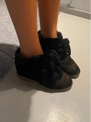 Isabel marant sneakers - Unika sneakers med kilklack i svart och brunt mocka-liknande material. Skorna har breda snören, detaljer i metall och en beige gummisula. Snygg design med högt skaft som ger extra edge till din outfit. Isabel maran Bobby