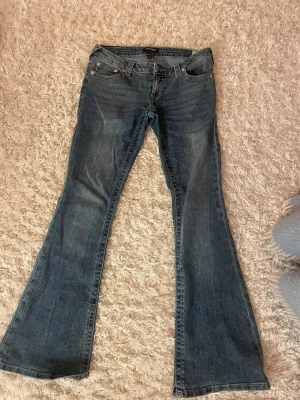 Low wasted bootcut jeans från True Religion - Säljer ett par blå bootcut jeans från True Religion med klassiska kontrastsömmar och broderade bakfickor. Jeansen har låg midja och är tillverkade i slitstark denim. Perfekta för dig som gillar en retro och avslappnad stil. Skriv om ni har frågor💕