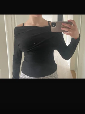 Svart offshoulder topp med lång ärm - Säljer en svart offshoulder topp med lång ärm och snygg drapering över bröstet. Toppen har en tight passform och är perfekt för dig som gillar stilrena plagg med en twist. Materialet känns mjukt och stretchigt, vilket gör den bekväm att bära.
