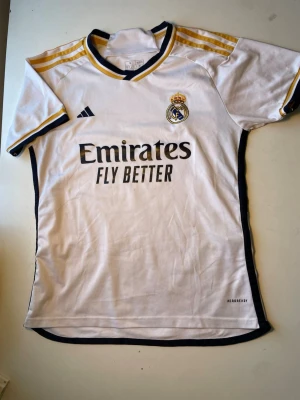 Real Madrid matchtröja Adidas vit - Säljer en vit Real Madrid matchtröja från Adidas med guldiga och svarta detaljer. Tröjan har klubbmärke och Adidas-logga på bröstet samt 'Emirates Fly Better' tryckt framtill. Tillverkad i lätt polyester med korta ärmar och sportig passform.