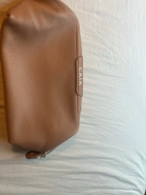 Brun rosa necessär från CAIA - Säljer en stilren brun rosa necessär från CAIA i konstläder med präglat mönster. Väskan har en avlång form, dragkedja upptill och diskret logga på sidan. Perfekt för smink eller småprylar när du är på språng.