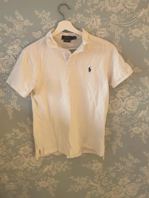 Vit pikétröja från Polo Ralph Lauren - Klassisk vit pikétröja från Polo Ralph Lauren med broderad logga på bröstet. Modellen är custom slim fit med korta ärmar och krage. Perfekt för en clean och stilren look.