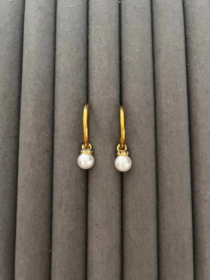Guldiga örhängen med pärlor - Snygga örhängen i guldfärgad metall med små vita pärlor som hänger i runda hoops. Klassisk och stilren design som passar till många olika looks. Perfekt för dig som gillar enkla men eleganta smycken.