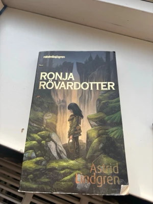 Ronja Rövardotter av Astrid Lindgren - En klassisk skönlitterär bok, 'Ronja Rövardotter' av Astrid Lindgren. Omslaget har en mörk, sagolik illustration med gröna och bruna toner, där Ronja står i en mystisk skogsmiljö. Perfekt för dig som gillar äventyr och fantasy.