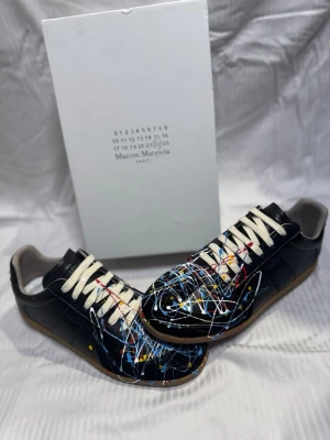 Maison Margiela  - Unika svarta sneakers från Maison Margiela med färgstänk i blått, vitt, gult och rött. Skorna har klassisk låg siluett, vita snören och beige sula. Tillverkade i skinn för en lyxig känsla och cool look.