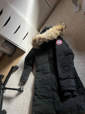 Svart lång dunparkas Canada Goose - Snygg svart lång parkas från Canada Goose med ikonisk patch på ärmen och stor huva med äkta päls. Jackan har flera fickor, dragkedja och knappar framtill samt justerbara muddar. Perfekt för kalla vinterdagar och riktigt varm.