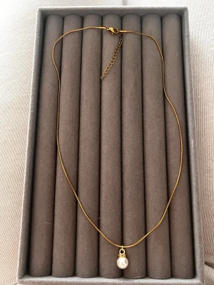 Guldhalsband med pärlhänge - Snyggt halsband i guldfärgad metall med en enkel kedja och ett litet vitt pärlhänge. Halsbandet har justerbar längd med förlängningskedja och karbinlås. Perfekt för dig som gillar minimalistisk och elegant stil.