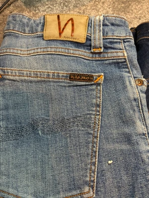 Ljusblå jeans från Nudie Jeans - Säljer ett par ljusblå jeans från Nudie Jeans med klassisk femficksmodell och snygga kontrastsömmar. Jeansen är tillverkade av 100% ekologisk bomull och har den ikoniska Nudie-loggan på bakfickan samt patch i läder bak i midjan.