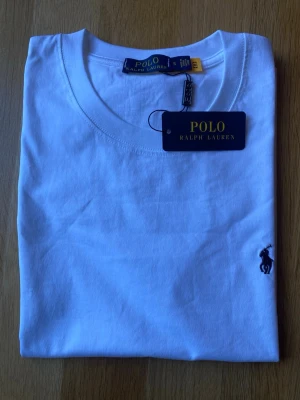 Vit t-shirt från Polo Ralph Lauren - Klassisk vit t-shirt från Polo Ralph Lauren med rund halsringning och korta ärmar. Tillverkad i mjuk bomull och prydd med den ikoniska broderade loggan i mörkblått på bröstet. Perfekt basplagg för en clean och stilren look.