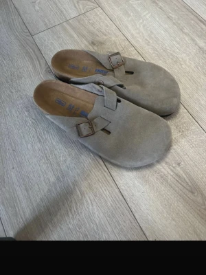 Birkenstock Boston, helt nya, stl 44 - Säljer ett par Birkenstock Boston tofflor i beige mocka med justerbart spänne i metall. Tofflorna har en ergonomisk fotbädd och öppen häl för skön komfort. Tofflorna är helt nya och aldrig använda. Nypris 1800kr. Pris kan diskuteras 