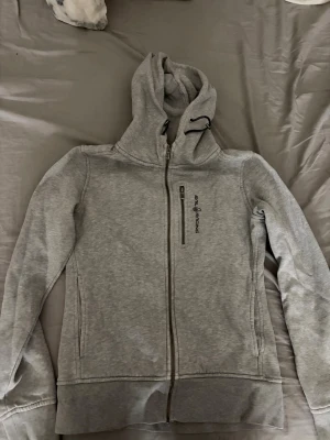 Grå zip-hoodie från Sail Racing - Snygg grå hoodie med dragkedja från Sail Racing. I storlek S. Tröjan har en praktisk huva med dragsko, bröstficka med dragkedja och diskret logga på framsidan. Tillverkad i mjukt sweatshirtmaterial som är perfekt för chill dagar.