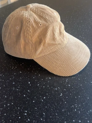 Beige manchesterkeps från Far Afield - Säljer en beige keps i manchestertyg från Far Afield. Klassisk böjd skärm och ventilerande öljetter på sidorna. Kepsen har en avslappnad passform och är tillverkad i mjukt, ribbat material som ger en retrovibe. Ordinarie pris: 550-600kr