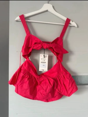 Rosa croppad linnetopp med rosett - Trendig rosa croppad linnetopp med breda axelband och stor rosett framtill. Toppen har öppen rygg och volangdetaljer nedtill för en lekfull look. Perfekt för varma dagar och sommarens alla äventyr.