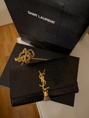Saint Laurent Kate Medium i croc-embossed läder - Saint Laurent Kate Medium i svart croc-embossed läder med gulddetaljer och klassisk YSL-logo. Köpt i Rom 2022, mycket fint skick och sparsamt använd. Tidlös modell som fortfarande säljs i butik (nypris ca 30 000 kr).  Mått: ca 24 x 15 x 5 cm. Kedjerem ca 48 cm drop.  Pris: 21 000 kr.  Finns i Stockholm, kan skickas.