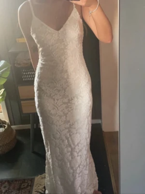 Vit långklänning med spetsdetaljer - Vit långklänning med smala axelband och v-ringning. Klänningen har fint blommigt mönster och öppen rygg med snörning. Perfekt för val och liknande. Figurnära passform och lätt material som ger en lyxig känsla.