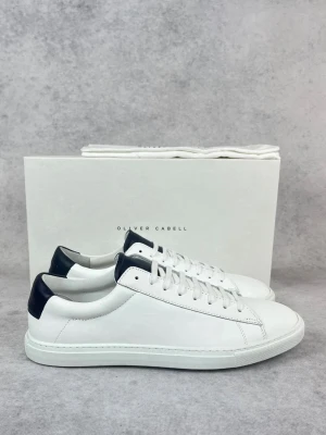 Vita sneakers från Oliver Cabell - Stilrena vita sneakers från Oliver Cabell med marinblå detalj på hälen och guldfärgad logga. Skorna har klassisk låg profil, rund tå och snörning. Tillverkade i skinn för en clean och tidlös look.