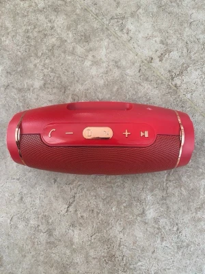 Röd bärbar högtalare med guldknappar - Snygg röd bärbar högtalare från HJ med guldfärgade knappar för volym, play/pause och samtal. Högtalaren har rundad form och metallgaller framtill. Perfekt för att ta med sig och spela musik trådlöst via Bluetooth.