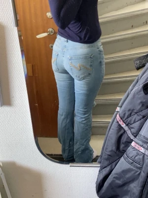 Ljusblå slitna bootcut jeans - Säljer ett par ljusblå bootcut jeans med slitningar över knäna och snygg broderad detalj på bakfickorna. Jeansen har klassisk femficksdesign och är tillverkade i denim med lite stretch för skön passform. Perfekta för en avslappnad och trendig look.