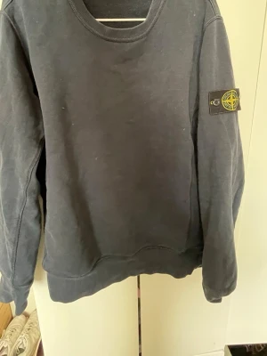 Blå sweatshirt från Stone Island - Mörkblå sweatshirt från Stone Island med klassisk rund halsringning och ikonisk patch på vänster ärm. Tröjan har ribbade muddar vid ärmslut och nederkant.  Skriv privat för fler bilder eller vidare frågor. Pris kan diskuteras vid en snabb affär! Såklart är den 100% äkta.