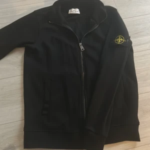 Svart kofta  från Stone Island - Säljer en stilren svart kofta  från Stone Island med dragkedja och ikonisk logga på ärmen. Jackan har en hög krage och långa ärmar, perfekt för en cool och avslappnad stil. ( Pris kan diskuteras)