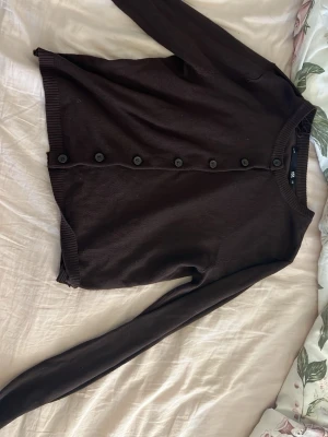 Mörkbrun kofta från H&M - En enkel och stilren mörkbrun kofta från H&M med långa ärmar och rund halsringning. Koftan har knappar hela vägen framtill och ribbade muddar vid ärmslut och nederkant. Perfekt att slänga över en t-shirt eller linne.