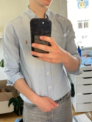 Ljusblå skjorta från Ralph Lauren - Klassisk ljusblå skjorta från Ralph Lauren med button-down krage och broderad logga på bröstet. Skjortan har lång ärm, vita knappar och är tillverkad i bomull. Perfekt för en clean och stilren look.
