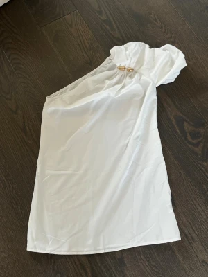 Vit oneshoulder klänning med gulddetalj - Säljer en vit kortklänning i oneshoulder-modell med snygg guldfärgad detalj vid axeln. Klänningen har en elegant och enkel design, perfekt för dig som vill sticka ut. Materialet känns mjukt och har ett fint fall.