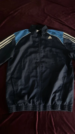 Adidas marinblå vindjacka med byxor - Snygg marinblå vindjacka från Adidas med ljusblå axelpartier och vita klassiska ränder längs ärmarna. Jackan har hel dragkedja, ståkrage och elastiska muddar. Tillverkad i lätt och funktionellt material, perfekt för blåsiga dagar. Byxorna är också tillverkade i lätt och funktionellt material och passar jätte bra för komfortläge.