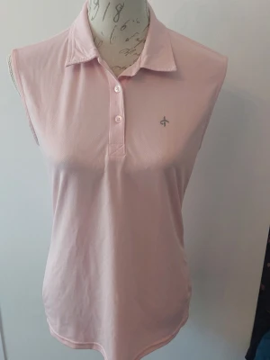 Ljusrosa ärmlös pikétopp Cross - Snygg ljusrosa ärmlös pikétopp från Cross med klassisk krage och tre knappar framtill. Tillverkad i mjukt material som känns skönt mot huden. Liten logga på bröstet och i nacken. 